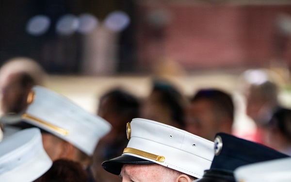 The image shows several uniformed members of the FDNY (Fire Department City of New York) seated at what appears to be a formal ceremony or memorial event. The central figure is an older man with a white beard wearing a white-topped dress uniform cap and a dark navy uniform adorned with service ribbons, medals, and an EMS star-of-life pin. Multiple FDNY patches and insignia are clearly visible on the uniforms.