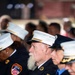 The image shows several uniformed members of the FDNY (Fire Department City of New York) seated at what appears to be a formal ceremony or memorial event. The central figure is an older man with a white beard wearing a white-topped dress uniform cap and a dark navy uniform adorned with service ribbons, medals, and an EMS star-of-life pin. Multiple FDNY patches and insignia are clearly visible on the uniforms.