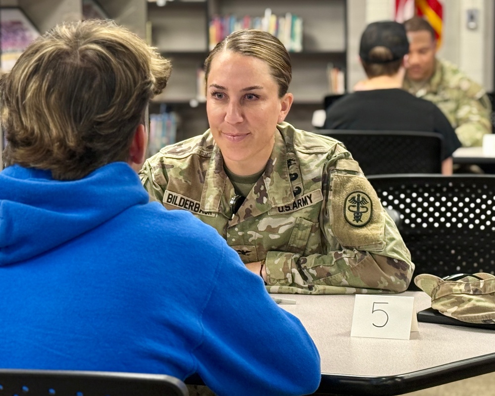 A U.S. Army soldier named Bilderbach, wearing OCP (Operational Camouflage Pattern) uniform with a Medical Corps insignia patch, is seated at table number 5 engaged in a one-on-one conversation with a civilian in a blue hoodie. The setting appears to be a school library or community center, suggesting a recruitment, career fair, or community outreach event. In the background, another soldier in OCP uniform is visible conversing with another civilian.