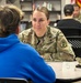A U.S. Army soldier named Bilderbach, wearing OCP (Operational Camouflage Pattern) uniform with a Medical Corps insignia patch, is seated at table number 5 engaged in a one-on-one conversation with a civilian in a blue hoodie. The setting appears to be a school library or community center, suggesting a recruitment, career fair, or community outreach event. In the background, another soldier in OCP uniform is visible conversing with another civilian.