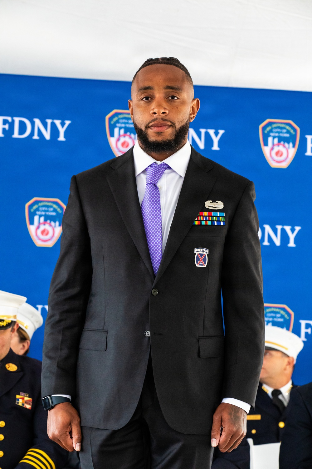 A man in a dark suit with a purple tie stands in front of an FDNY (Fire Department of New York) step-and-repeat banner backdrop. He is wearing military medals, ribbons, and a 10th Mountain Division patch on his lapel, suggesting he is a military veteran attending an official FDNY ceremony. FDNY officers in white dress caps are partially visible in the background.