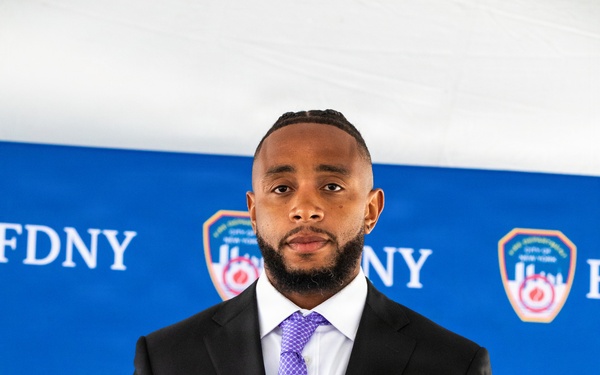 A man in a dark suit with a purple tie stands in front of an FDNY (Fire Department of New York) step-and-repeat banner backdrop. He is wearing military medals, ribbons, and a 10th Mountain Division patch on his lapel, suggesting he is a military veteran attending an official FDNY ceremony. FDNY officers in white dress caps are partially visible in the background.