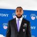 A man in a dark suit with a purple tie stands in front of an FDNY (Fire Department of New York) step-and-repeat banner backdrop. He is wearing military medals, ribbons, and a 10th Mountain Division patch on his lapel, suggesting he is a military veteran attending an official FDNY ceremony. FDNY officers in white dress caps are partially visible in the background.