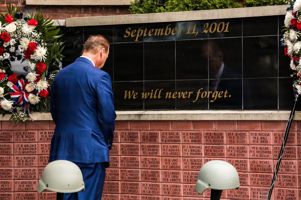 A man in a blue suit stands before a September 11, 2001 memorial wall bearing the inscription 'We will never forget.' The black granite memorial panel is flanked by red, white, and blue floral wreaths, and the brick wall below contains engraved names of victims or first responders. Two military helmets are visible in the foreground, suggesting a formal commemorative ceremony.