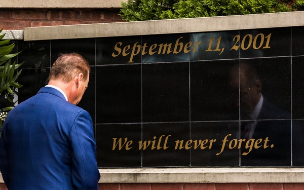 A man in a blue suit stands before a September 11, 2001 memorial wall bearing the inscription 'We will never forget.' The black granite memorial panel is flanked by red, white, and blue floral wreaths, and the brick wall below contains engraved names of victims or first responders. Two military helmets are visible in the foreground, suggesting a formal commemorative ceremony.