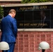 A man in a blue suit stands before a September 11, 2001 memorial wall bearing the inscription 'We will never forget.' The black granite memorial panel is flanked by red, white, and blue floral wreaths, and the brick wall below contains engraved names of victims or first responders. Two military helmets are visible in the foreground, suggesting a formal commemorative ceremony.