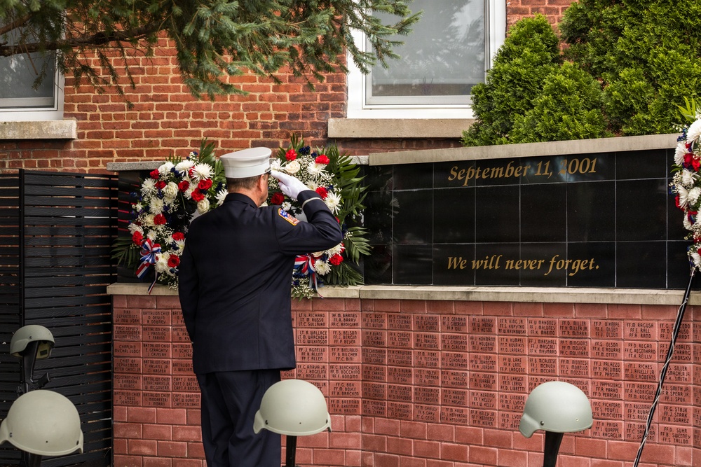 A uniformed firefighter or fire official in dress blues and white cap is saluting a September 11, 2001 memorial wall inscribed with 'We will never forget.' The memorial features a black granite panel surrounded by brick walls engraved with names of fallen first responders. Ceremonial wreaths with red, white, and blue flowers flank the memorial, and firefighter helmets are placed in the foreground.