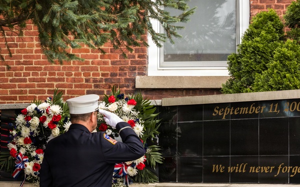 A uniformed firefighter or fire official in dress blues and white cap is saluting a September 11, 2001 memorial wall inscribed with 'We will never forget.' The memorial features a black granite panel surrounded by brick walls engraved with names of fallen first responders. Ceremonial wreaths with red, white, and blue flowers flank the memorial, and firefighter helmets are placed in the foreground.