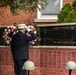 A uniformed firefighter or fire official in dress blues and white cap is saluting a September 11, 2001 memorial wall inscribed with 'We will never forget.' The memorial features a black granite panel surrounded by brick walls engraved with names of fallen first responders. Ceremonial wreaths with red, white, and blue flowers flank the memorial, and firefighter helmets are placed in the foreground.