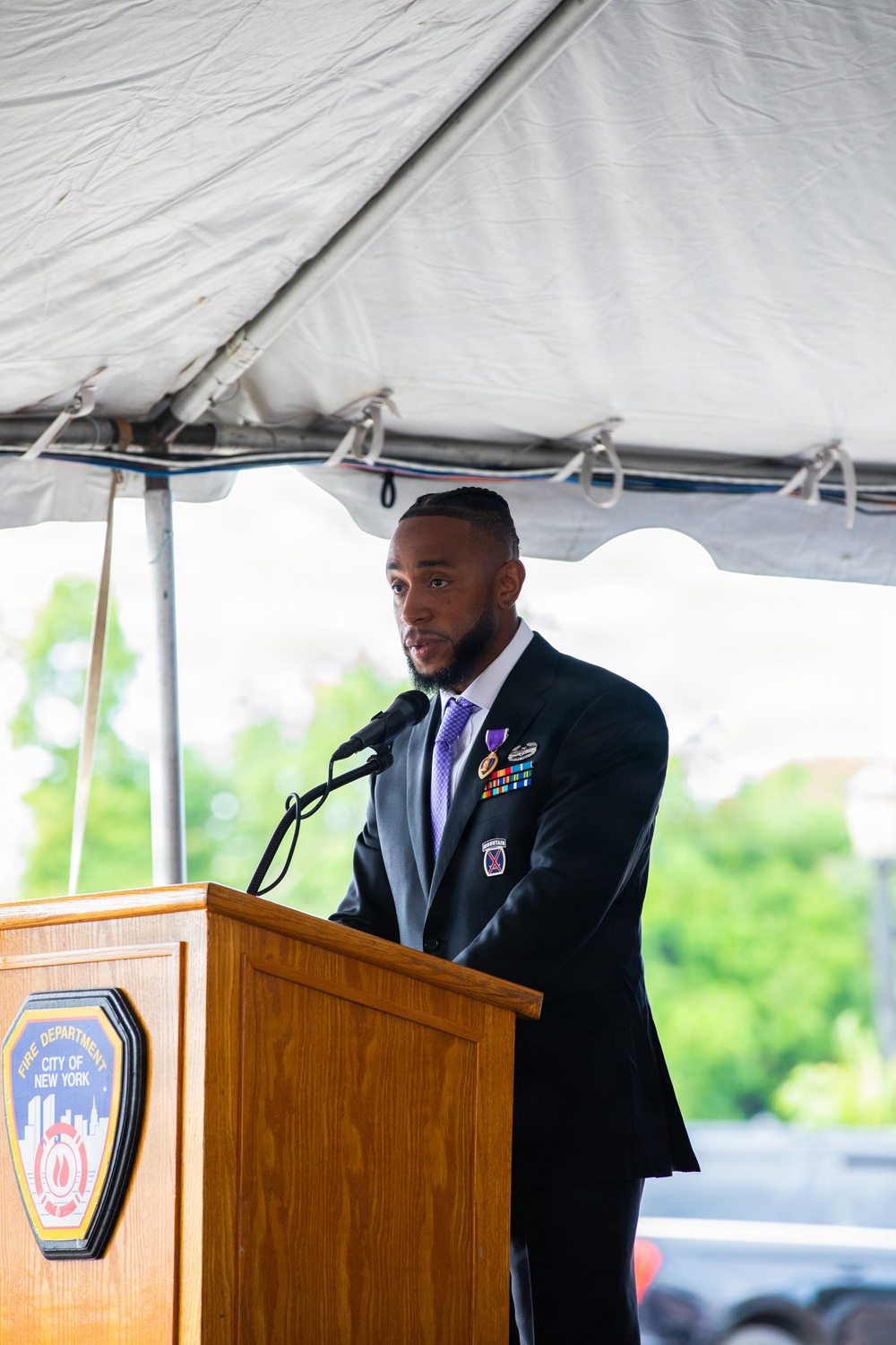 A man in a dark suit is speaking at a podium bearing the Fire Department City of New York (FDNY) seal, under a white event tent. He is wearing a purple medal (consistent with a Purple Heart) along with military service ribbons on his lapel, and a unit patch on his sleeve. The setting appears to be an outdoor formal ceremony.