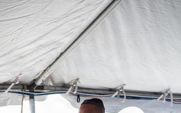 A man in a dark suit is speaking at a podium bearing the Fire Department City of New York (FDNY) seal, under a white event tent. He is wearing a purple medal (consistent with a Purple Heart) along with military service ribbons on his lapel, and a unit patch on his sleeve. The setting appears to be an outdoor formal ceremony.