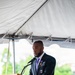 A man in a dark suit is speaking at a podium bearing the Fire Department City of New York (FDNY) seal, under a white event tent. He is wearing a purple medal (consistent with a Purple Heart) along with military service ribbons on his lapel, and a unit patch on his sleeve. The setting appears to be an outdoor formal ceremony.