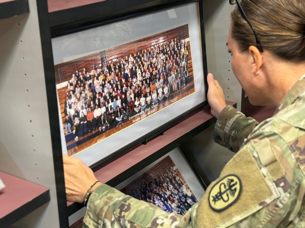 A person in U.S. Army camouflage (OCP/MultiCam) uniform is examining a framed panoramic class photograph labeled 'Leavenworth High School Senior Class of 2001' on a storage shelf. The soldier appears to be browsing through multiple framed class photos stored on shelving units. The patch on the uniform sleeve appears to be a unit insignia.