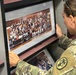 A person in U.S. Army camouflage (OCP/MultiCam) uniform is examining a framed panoramic class photograph labeled 'Leavenworth High School Senior Class of 2001' on a storage shelf. The soldier appears to be browsing through multiple framed class photos stored on shelving units. The patch on the uniform sleeve appears to be a unit insignia.