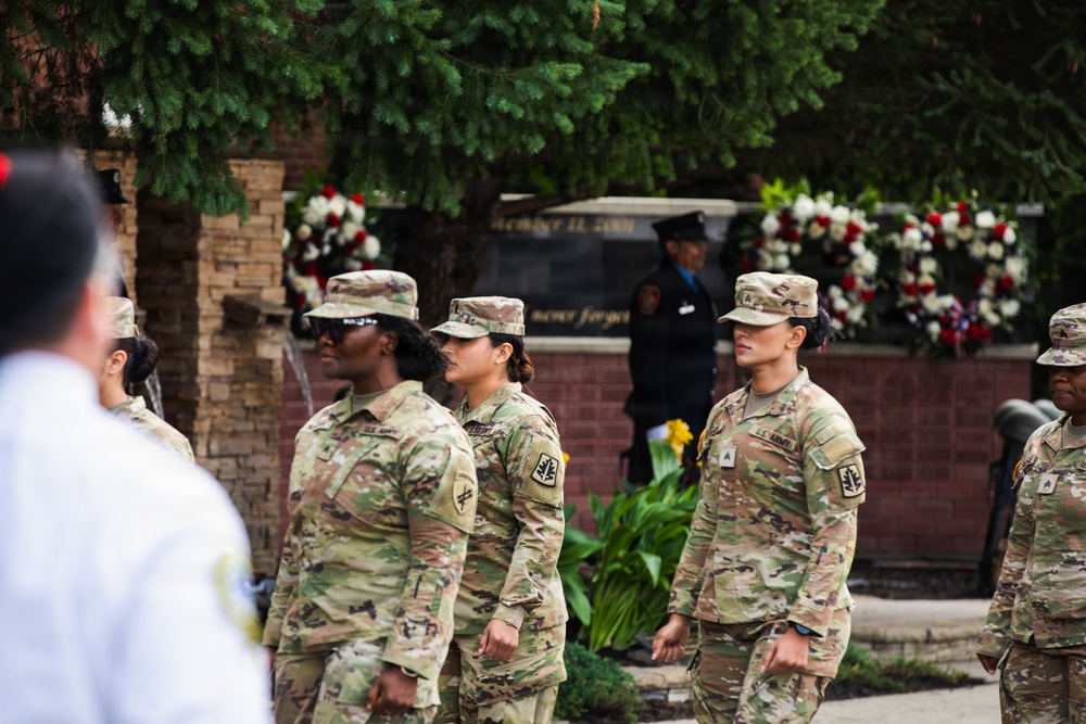 A group of U.S. Army soldiers in OCP (Operational Camouflage Pattern) uniforms are participating in a formal military ceremony, likely a memorial or commemoration event. In the background, a monument or memorial wall is visible, adorned with red, white, and floral wreaths. The setting and decorations suggest this may be a September 11 memorial or similar patriotic observance.
