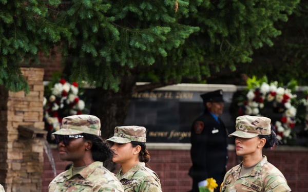 A group of U.S. Army soldiers in OCP (Operational Camouflage Pattern) uniforms are participating in a formal military ceremony, likely a memorial or commemoration event. In the background, a monument or memorial wall is visible, adorned with red, white, and floral wreaths. The setting and decorations suggest this may be a September 11 memorial or similar patriotic observance.