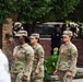 A group of U.S. Army soldiers in OCP (Operational Camouflage Pattern) uniforms are participating in a formal military ceremony, likely a memorial or commemoration event. In the background, a monument or memorial wall is visible, adorned with red, white, and floral wreaths. The setting and decorations suggest this may be a September 11 memorial or similar patriotic observance.