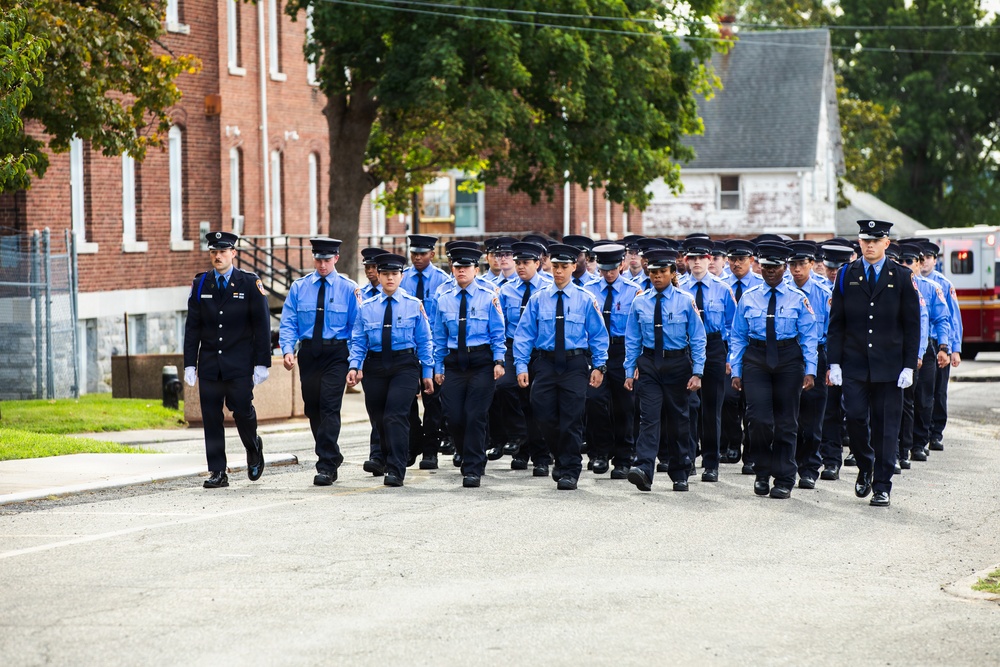 A large group of uniformed personnel, likely fire academy recruits or cadets, are marching in formation along a paved road. They are wearing light blue shirts, black trousers, and black uniform caps, accompanied by two supervisors in darker navy uniforms on the flanks. The setting appears to be a training facility or institutional campus with brick buildings and trees in the background.