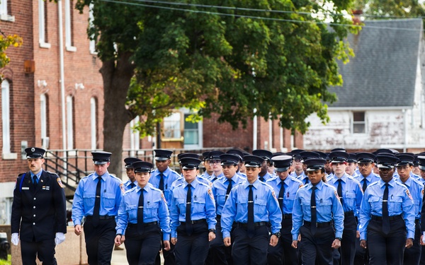 A large group of uniformed personnel, likely fire academy recruits or cadets, are marching in formation along a paved road. They are wearing light blue shirts, black trousers, and black uniform caps, accompanied by two supervisors in darker navy uniforms on the flanks. The setting appears to be a training facility or institutional campus with brick buildings and trees in the background.