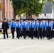 A large group of uniformed personnel, likely fire academy recruits or cadets, are marching in formation along a paved road. They are wearing light blue shirts, black trousers, and black uniform caps, accompanied by two supervisors in darker navy uniforms on the flanks. The setting appears to be a training facility or institutional campus with brick buildings and trees in the background.