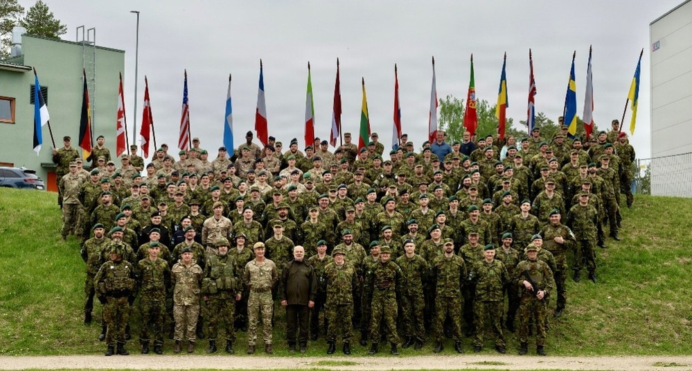 A large multinational military group photo featuring approximately 150-200 personnel in various military uniforms and camouflage patterns. Multiple national flags are displayed in the background, indicating a NATO or multinational coalition gathering. The photo appears to be taken at a military installation or base, likely in the Baltic region based on visible flags.