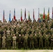 A large multinational military group photo featuring approximately 150-200 personnel in various military uniforms and camouflage patterns. Multiple national flags are displayed in the background, indicating a NATO or multinational coalition gathering. The photo appears to be taken at a military installation or base, likely in the Baltic region based on visible flags.