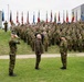 A large military formation ceremony is taking place outdoors, with hundreds of soldiers in camouflage uniforms assembled in rows behind multiple national flags. In the foreground, two uniformed soldiers are saluting a civilian figure dressed in olive/dark clothing, suggesting an official review or greeting ceremony. The presence of numerous national flags indicates this is a multinational NATO or allied forces gathering.