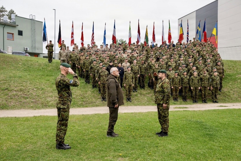 A large military formation ceremony is taking place outdoors, with hundreds of soldiers in camouflage uniforms assembled in rows behind multiple national flags. In the foreground, two uniformed soldiers are saluting a civilian figure dressed in olive/dark clothing, suggesting an official review or greeting ceremony. The presence of numerous national flags indicates this is a multinational NATO or allied forces gathering.