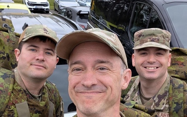 Three military personnel in camouflage uniforms are taking a selfie in what appears to be a parking lot. The person in the center is taking the photo and wearing a patch that reads 'Intelligence Squadron' on their uniform. They appear to be in good spirits, smiling for the camera.