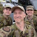 Three military personnel in camouflage uniforms are taking a selfie in what appears to be a parking lot. The person in the center is taking the photo and wearing a patch that reads 'Intelligence Squadron' on their uniform. They appear to be in good spirits, smiling for the camera.