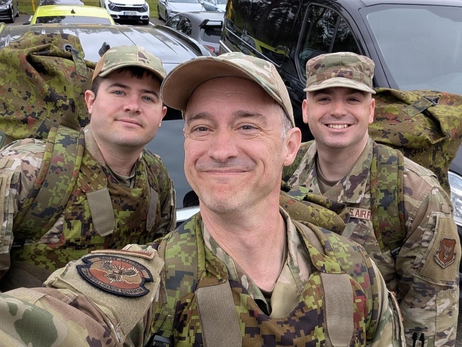 Three military personnel in camouflage uniforms are taking a selfie in what appears to be a parking lot. The person in the center is taking the photo and wearing a patch that reads 'Intelligence Squadron' on their uniform. They appear to be in good spirits, smiling for the camera.