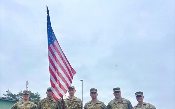 Six U.S. Army soldiers in Operational Camouflage Pattern (OCP) uniforms stand in a group pose on a grass field. One soldier in the center is holding a U.S. national flag on a staff. The setting appears to be a military installation, likely during a formal ceremony or event.