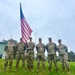 Six U.S. Army soldiers in Operational Camouflage Pattern (OCP) uniforms stand in a group pose on a grass field. One soldier in the center is holding a U.S. national flag on a staff. The setting appears to be a military installation, likely during a formal ceremony or event.