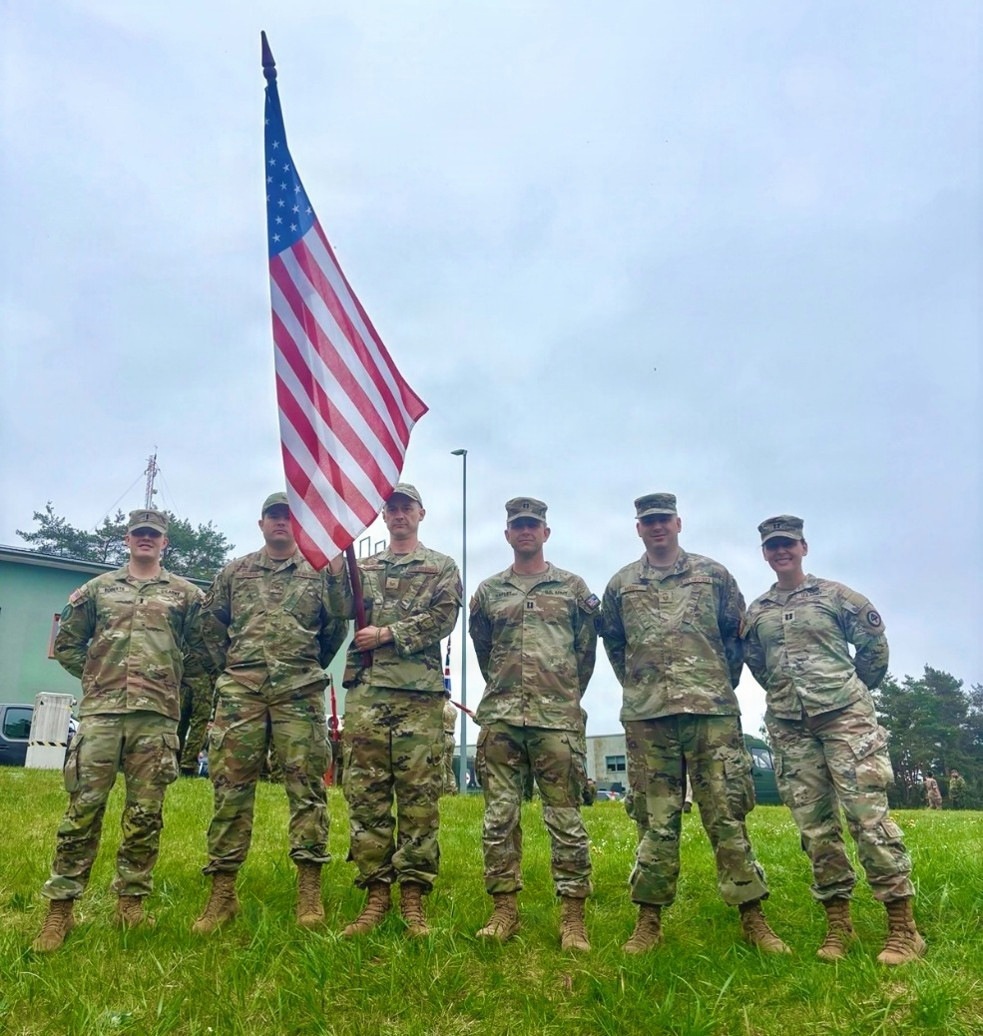 Six U.S. Army soldiers in Operational Camouflage Pattern (OCP) uniforms stand in a group pose on a grass field. One soldier in the center is holding a U.S. national flag on a staff. The setting appears to be a military installation, likely during a formal ceremony or event.