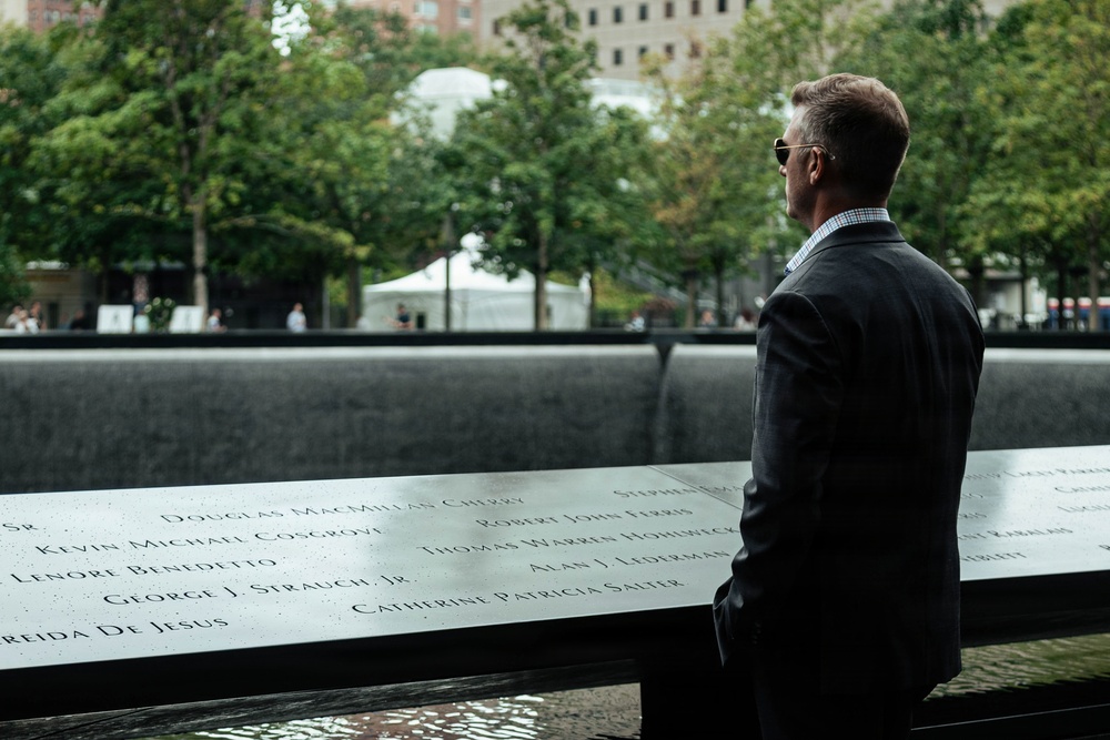 A man in a dark suit and sunglasses stands solemnly at the 9/11 Memorial in New York City, viewed from behind. He is looking out over one of the memorial's reflecting pools, surrounded by the bronze parapets engraved with victims' names. The setting is outdoors with trees and urban buildings visible in the background.