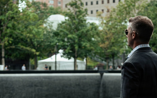 A man in a dark suit and sunglasses stands solemnly at the 9/11 Memorial in New York City, viewed from behind. He is looking out over one of the memorial's reflecting pools, surrounded by the bronze parapets engraved with victims' names. The setting is outdoors with trees and urban buildings visible in the background.