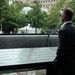 A man in a dark suit and sunglasses stands solemnly at the 9/11 Memorial in New York City, viewed from behind. He is looking out over one of the memorial's reflecting pools, surrounded by the bronze parapets engraved with victims' names. The setting is outdoors with trees and urban buildings visible in the background.