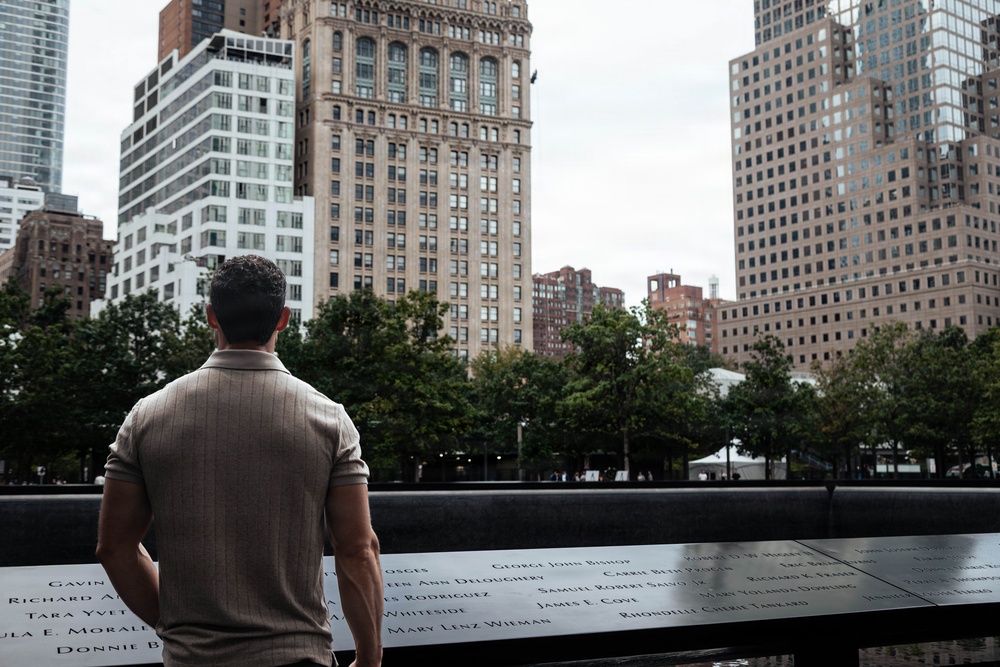 A man stands with his back to the camera at the 9/11 Memorial in Lower Manhattan, New York City, looking out over one of the memorial pools. The bronze parapet surrounding the pool is engraved with the names of victims. The scene is set against a backdrop of urban skyscrapers and trees under an overcast sky.