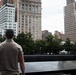 A man stands with his back to the camera at the 9/11 Memorial in Lower Manhattan, New York City, looking out over one of the memorial pools. The bronze parapet surrounding the pool is engraved with the names of victims. The scene is set against a backdrop of urban skyscrapers and trees under an overcast sky.