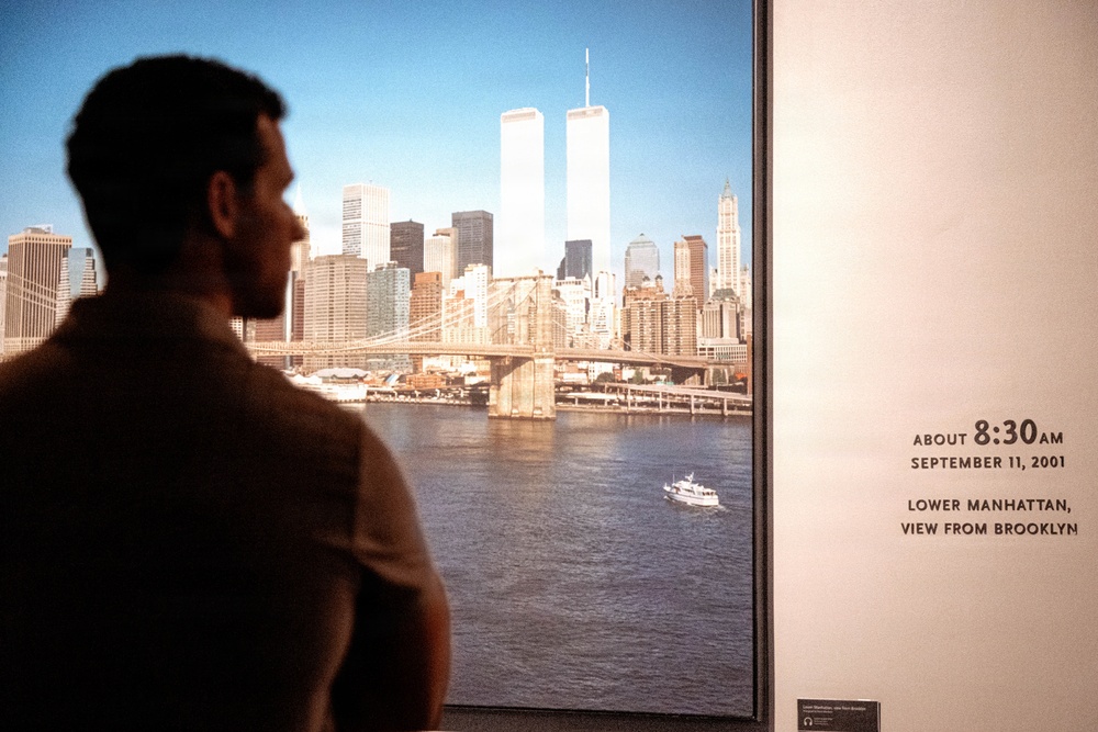 A person stands in silhouette viewing a large framed photograph displayed in what appears to be a museum or memorial exhibition. The photograph shows the Lower Manhattan skyline including the Twin Towers of the World Trade Center, taken from Brooklyn at approximately 8:30 AM on September 11, 2001 — roughly 46 minutes before the first plane struck. The wall text beside the image identifies the time, date, and vantage point of the photograph.