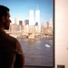 A person stands in silhouette viewing a large framed photograph displayed in what appears to be a museum or memorial exhibition. The photograph shows the Lower Manhattan skyline including the Twin Towers of the World Trade Center, taken from Brooklyn at approximately 8:30 AM on September 11, 2001 — roughly 46 minutes before the first plane struck. The wall text beside the image identifies the time, date, and vantage point of the photograph.