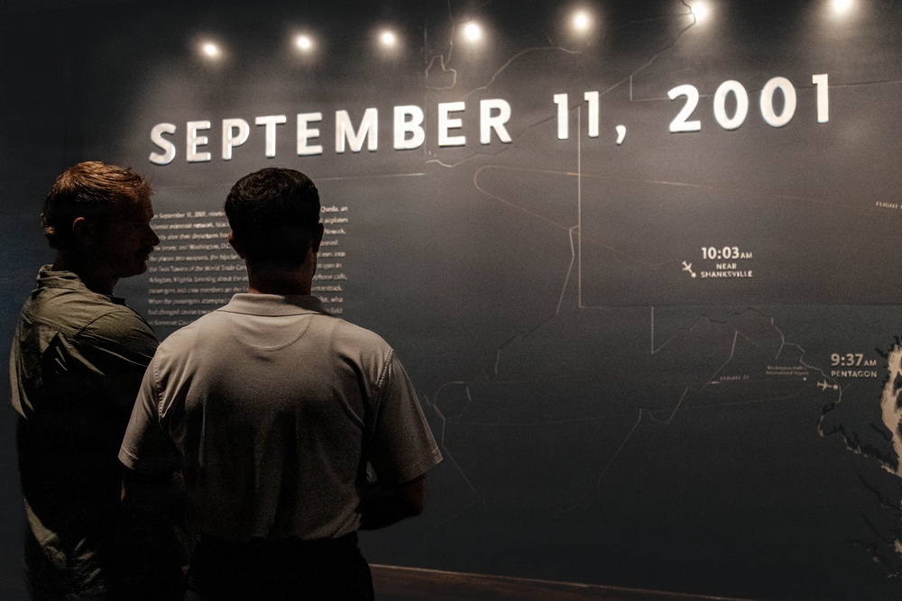 Two individuals are viewing a memorial or museum exhibit dedicated to the September 11, 2001 terrorist attacks. The display features a large dark wall with the date 'SEPTEMBER 11, 2001' in illuminated white text, along with a map showing flight paths and impact times. Visible timestamps and locations including '10:03 AM Near Shanksville' and '9:37 AM Pentagon' are marked on the exhibit.