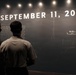 Two individuals are viewing a memorial or museum exhibit dedicated to the September 11, 2001 terrorist attacks. The display features a large dark wall with the date 'SEPTEMBER 11, 2001' in illuminated white text, along with a map showing flight paths and impact times. Visible timestamps and locations including '10:03 AM Near Shanksville' and '9:37 AM Pentagon' are marked on the exhibit.