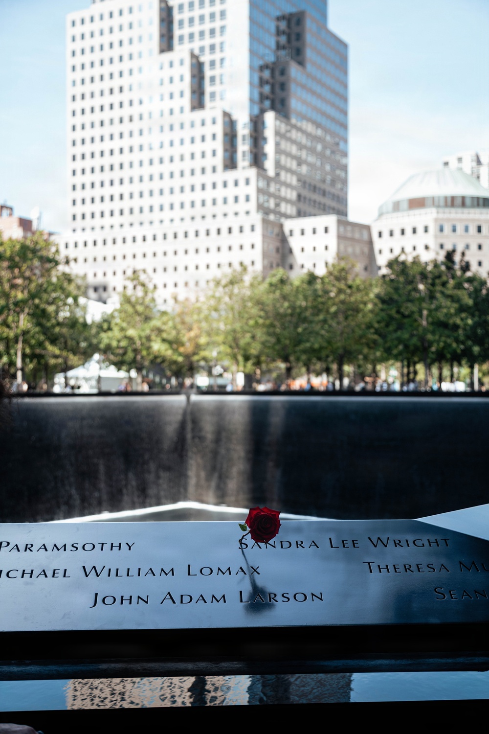 This image shows the 9/11 Memorial in Lower Manhattan, New York City, featuring one of the twin reflecting pools with engraved bronze parapets bearing victims' names. A single red rose has been placed on the parapet, a common tribute left by visitors and family members. The background shows surrounding trees and the Manhattan skyline with tall buildings under a clear sky.