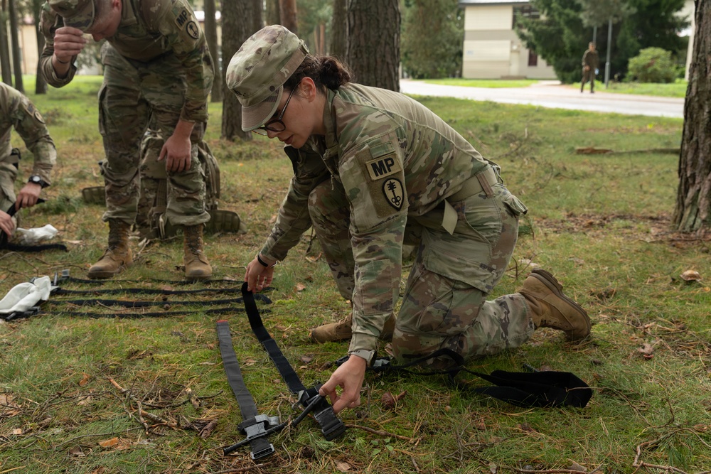 A U.S. Army Military Police (MP) soldier wearing OCP (Operational Camouflage Pattern) uniform is kneeling on grass, handling black straps or restraints on the ground. Additional MP soldiers are visible in the background performing similar tasks in what appears to be a training exercise. The setting is an outdoor wooded area on what appears to be a military installation.