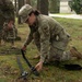 A U.S. Army Military Police (MP) soldier wearing OCP (Operational Camouflage Pattern) uniform is kneeling on grass, handling black straps or restraints on the ground. Additional MP soldiers are visible in the background performing similar tasks in what appears to be a training exercise. The setting is an outdoor wooded area on what appears to be a military installation.