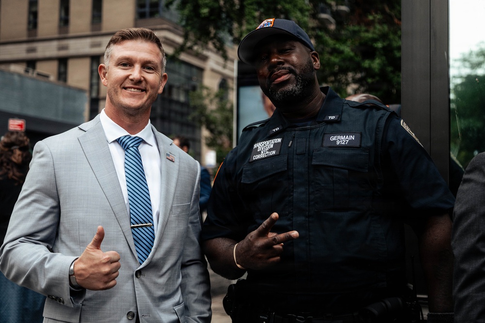 Two men pose together outdoors on an urban street. One man is dressed in a light gray suit with a blue striped tie, giving a thumbs-up gesture. The other is a uniformed officer from the NY Correction Transportation Division, identified by his nameplate as 'Germain 9127', making a peace sign gesture.
