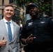 Two men pose together outdoors on an urban street. One man is dressed in a light gray suit with a blue striped tie, giving a thumbs-up gesture. The other is a uniformed officer from the NY Correction Transportation Division, identified by his nameplate as 'Germain 9127', making a peace sign gesture.