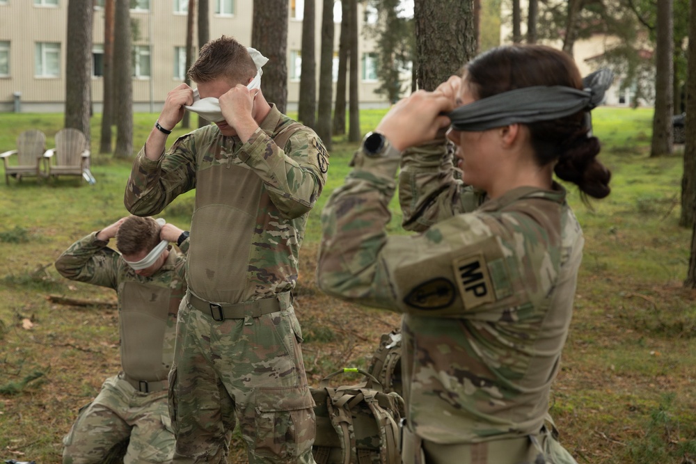 Three U.S. Army soldiers in OCP (Operational Camouflage Pattern) uniforms are engaged in what appears to be a field training exercise involving eye decontamination or blindfold procedures. The soldier on the right is visibly wearing an 'MP' (Military Police) patch and is adjusting a dark blindfold over her eyes. The setting appears to be a wooded area adjacent to a military installation or base.