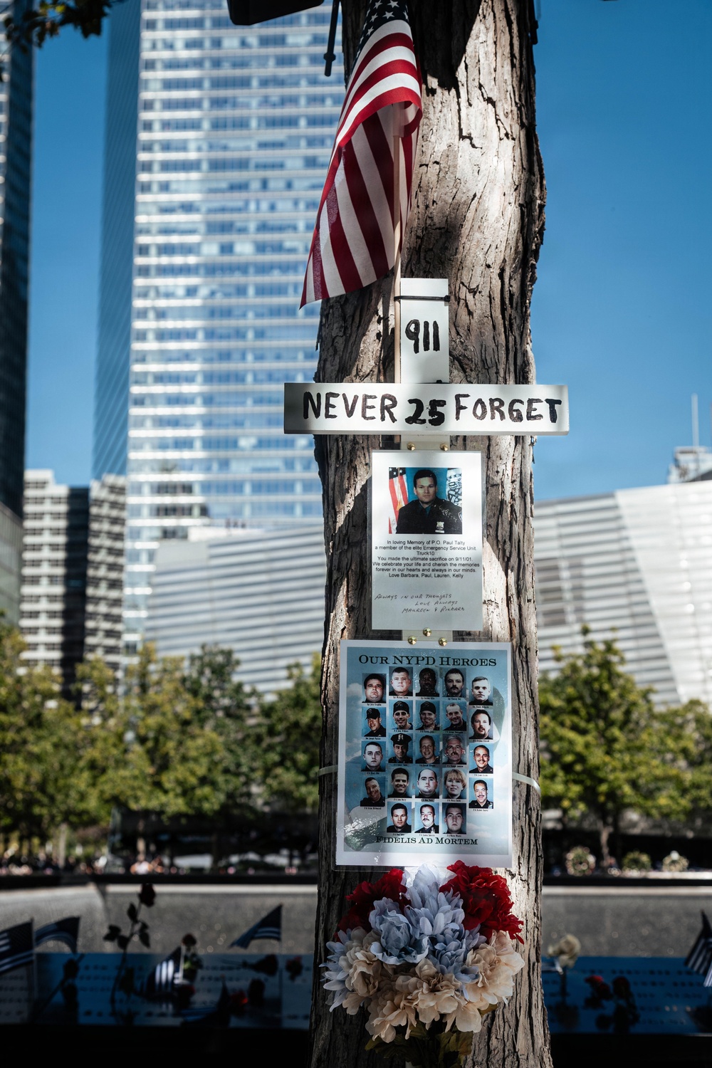A memorial tree at the 9/11 Memorial in New York City, decorated with tributes marking the 25th anniversary of the September 11 attacks. The tree bears a 'Never 25 Forget' sign, a memorial poster for P.O. Paul Talty of the NYPD Emergency Service Unit, and a group tribute sheet titled 'Our NYPD Heroes' featuring photos of fallen officers. Artificial flowers, an American flag, and a '911' placard are also attached to the tree.